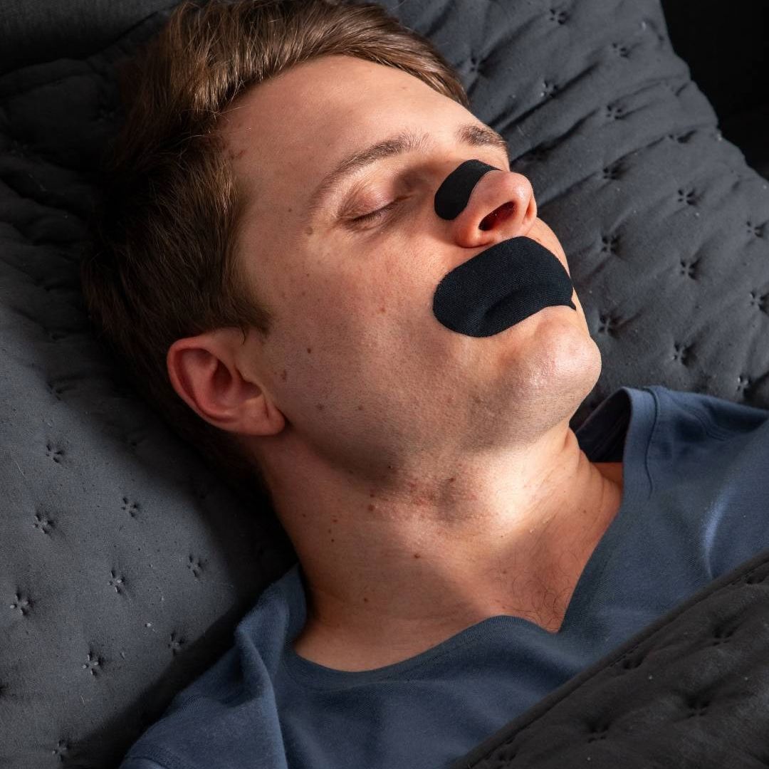 Man asleep on a dark pillow wearing a black Elite Sleep mouth tape strip across his lips.